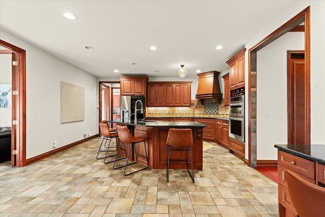 kitchen with an island with sink, stainless steel appliances, custom range hood, backsplash, and a breakfast bar area