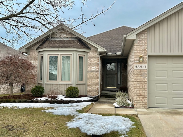 snow covered property entrance featuring board and batten siding, brick siding, an attached garage, and roof with shingles