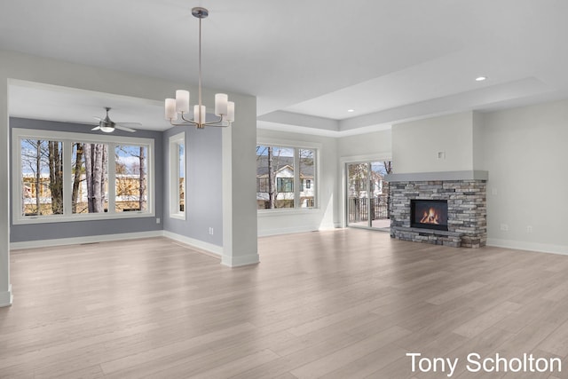 unfurnished living room featuring light wood-style floors, baseboards, a stone fireplace, a tray ceiling, and a notable chandelier