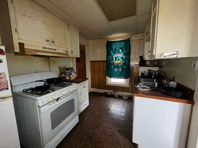 kitchen featuring a paneled ceiling, white range with gas cooktop, sink, wood walls, and refrigerator