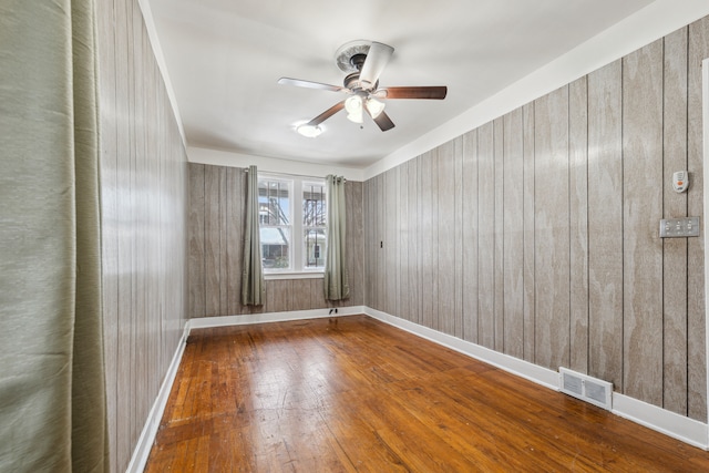 spare room with ceiling fan, wooden walls, wood finished floors, visible vents, and baseboards