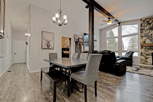 dining space featuring vaulted ceiling with beams, hardwood / wood-style flooring, a stone fireplace, a textured ceiling, and ceiling fan with notable chandelier