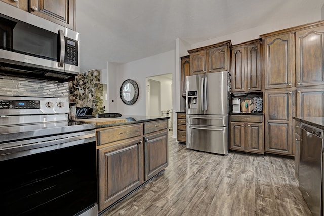 kitchen with stainless steel appliances, backsplash, dark brown cabinets, wood-type flooring, and dark stone counters