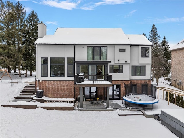 snow covered back of property with a trampoline, brick siding, and a chimney