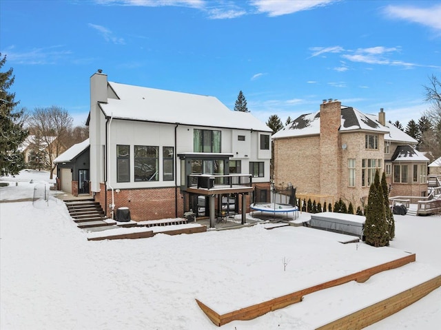 snow covered rear of property with a trampoline, a chimney, and brick siding