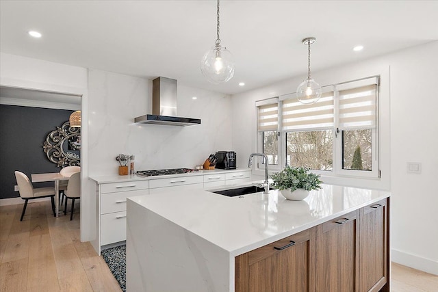 kitchen with light countertops, white cabinetry, a sink, an island with sink, and wall chimney exhaust hood