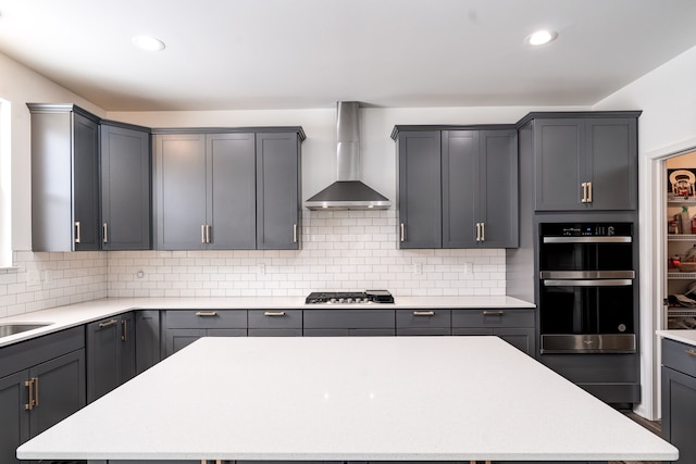 kitchen featuring a center island, wall chimney exhaust hood, tasteful backsplash, gray cabinets, and multiple ovens