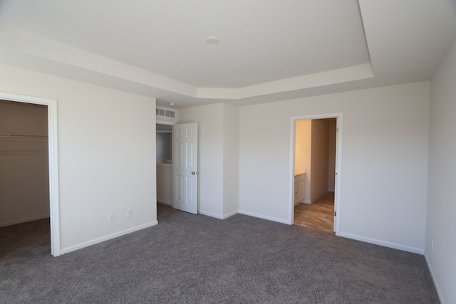 unfurnished bedroom with a spacious closet, ensuite bathroom, dark carpet, a tray ceiling, and a closet