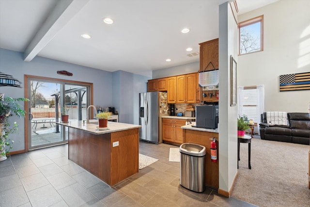 kitchen with a kitchen island with sink, decorative backsplash, beam ceiling, light colored carpet, and stainless steel fridge