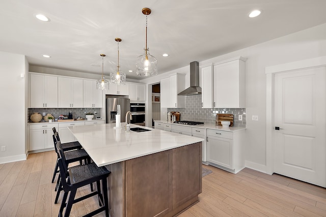 kitchen featuring stainless steel appliances, wall chimney exhaust hood, white cabinetry, and an island with sink