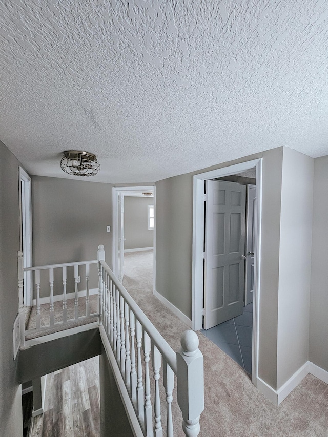 hallway featuring carpet floors, an upstairs landing, baseboards, and a textured ceiling