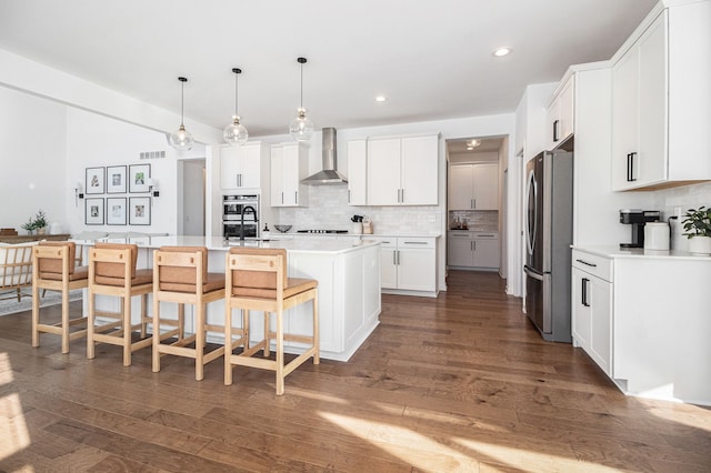 kitchen with wall chimney exhaust hood, white cabinetry, a center island with sink, appliances with stainless steel finishes, and decorative backsplash