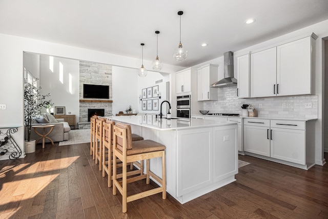 kitchen with wall chimney exhaust hood, white cabinetry, hanging light fixtures, an island with sink, and backsplash