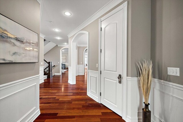 corridor featuring crown molding, a decorative wall, stairway, dark wood-type flooring, and wainscoting