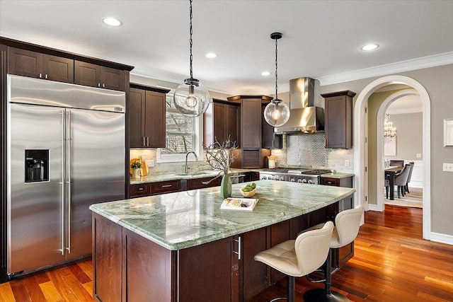 kitchen with arched walkways, stainless steel appliances, a sink, wall chimney range hood, and dark brown cabinets