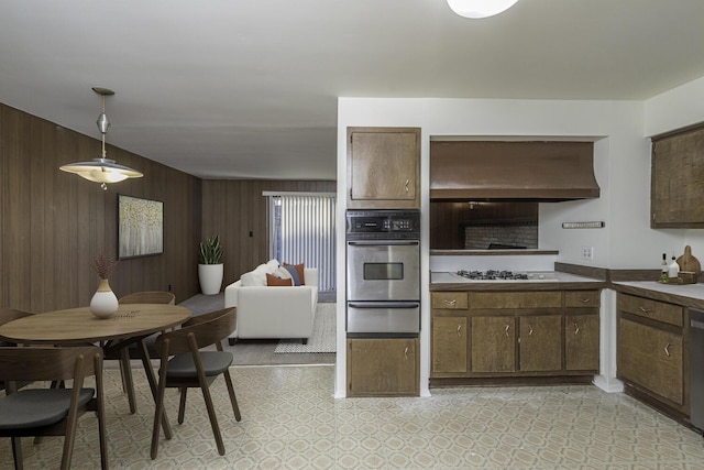 kitchen featuring dark brown cabinetry, decorative light fixtures, wall chimney range hood, stainless steel appliances, and wooden walls