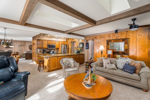 carpeted living room with beamed ceiling, ceiling fan, wooden walls, and sink