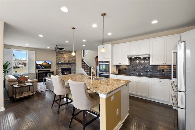 kitchen with sink, a stone fireplace, a kitchen island with sink, stainless steel appliances, and decorative light fixtures