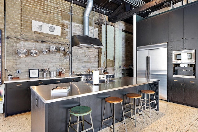 kitchen with brick wall, a towering ceiling, sink, stainless steel appliances, and a center island with sink