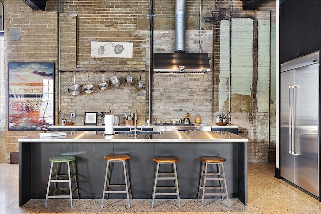 kitchen with wall chimney range hood, sink, a breakfast bar, stainless steel built in refrigerator, and brick wall