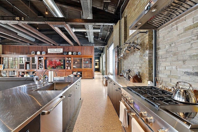 kitchen featuring wall chimney range hood, sink, stainless steel counters, stainless steel appliances, and brick wall