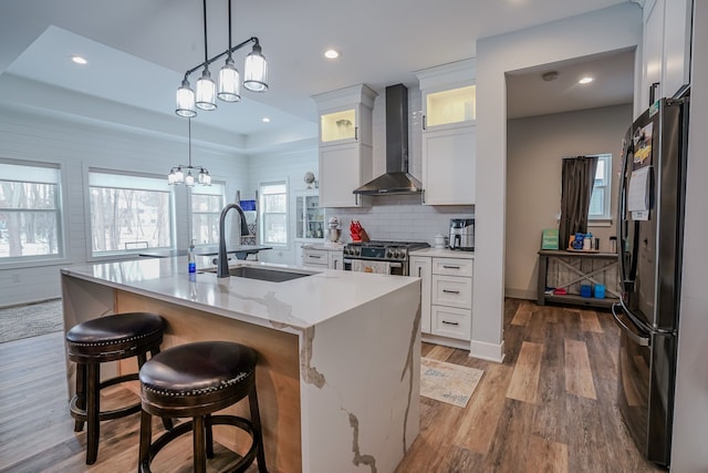 kitchen with white cabinetry, black refrigerator, stainless steel range with gas cooktop, a kitchen island with sink, and wall chimney range hood