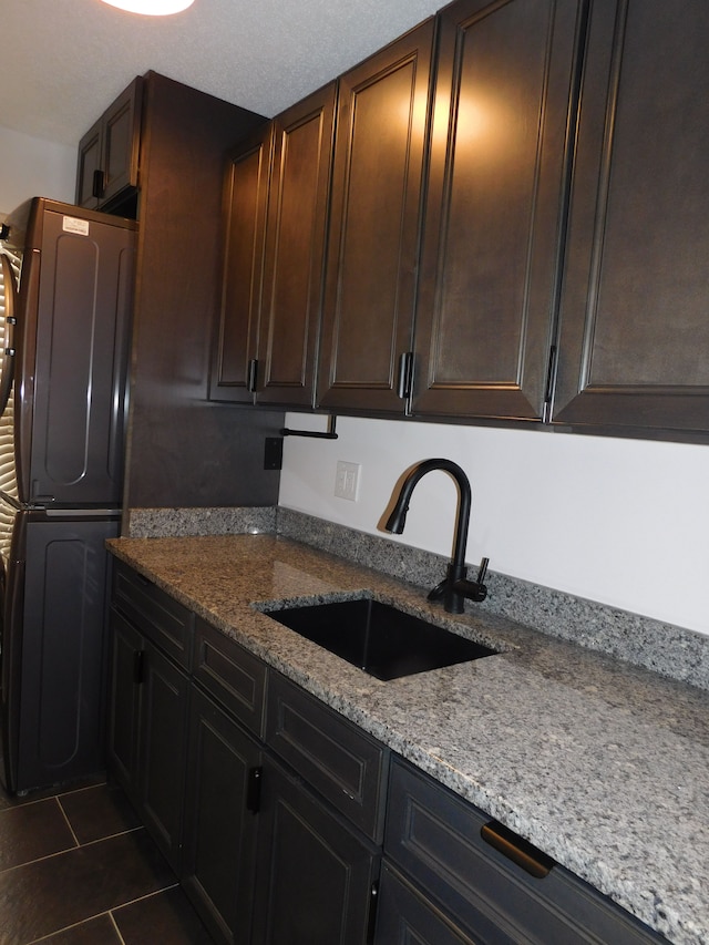 kitchen featuring stacked washer and dryer, sink, light stone counters, dark brown cabinetry, and dark tile patterned flooring