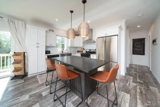 kitchen with wall chimney exhaust hood, decorative light fixtures, appliances with stainless steel finishes, a kitchen island, and white cabinets