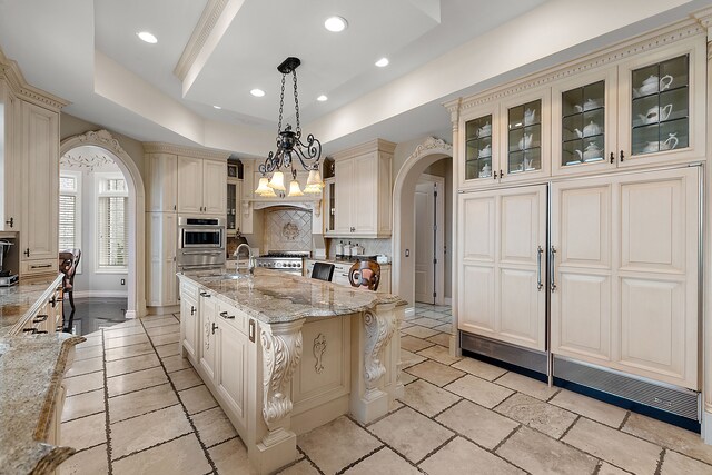 kitchen with light stone counters, a raised ceiling, an island with sink, pendant lighting, and cream cabinetry