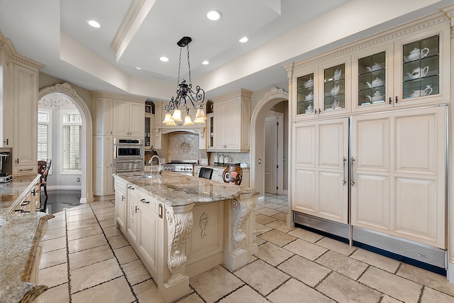 kitchen featuring arched walkways, a kitchen island with sink, hanging light fixtures, cream cabinetry, and glass insert cabinets