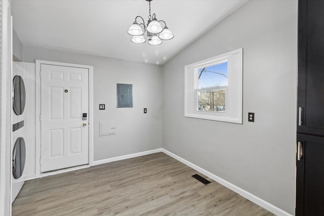 laundry room featuring an inviting chandelier, stacked washing maching and dryer, electric panel, and light hardwood / wood-style flooring