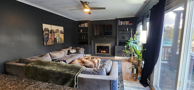 living room featuring ceiling fan, a fireplace, wood-type flooring, ornamental molding, and built in shelves