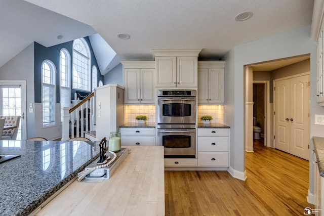 kitchen featuring vaulted ceiling, white cabinets, dark stone counters, stainless steel double oven, and light hardwood / wood-style flooring