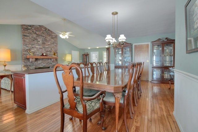 dining area featuring a brick fireplace, ceiling fan with notable chandelier, vaulted ceiling, and light wood-type flooring