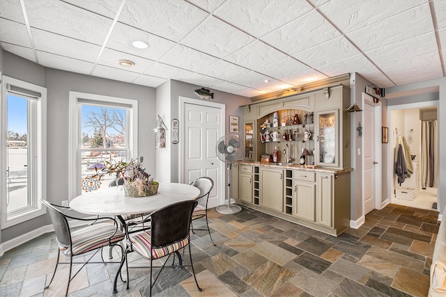 dining space featuring wet bar, stone tile flooring, a drop ceiling, and baseboards