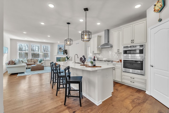 kitchen with wall chimney range hood, a kitchen bar, light wood-type flooring, decorative backsplash, and stainless steel appliances