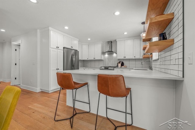 kitchen with white cabinetry, kitchen peninsula, stainless steel refrigerator, and wall chimney exhaust hood