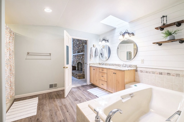 bathroom with tiled tub, hardwood / wood-style flooring, vanity, a stone fireplace, and vaulted ceiling