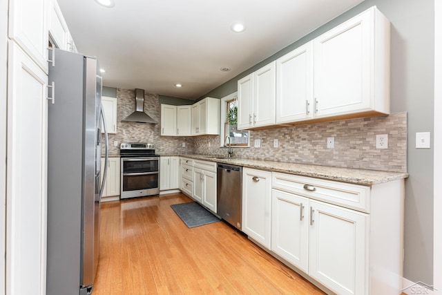 kitchen with wall chimney range hood, stainless steel appliances, light stone countertops, light hardwood / wood-style floors, and white cabinets