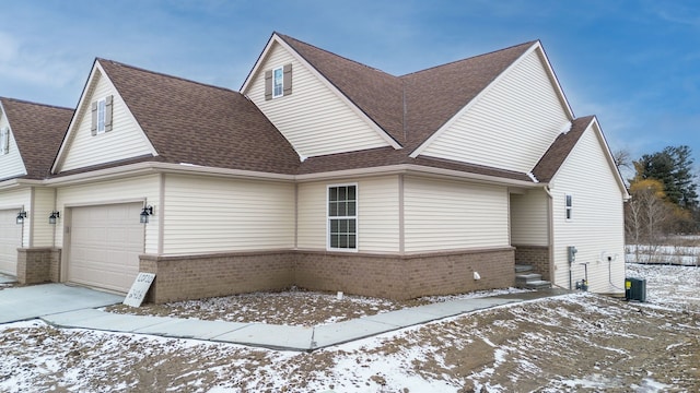 view of snowy exterior with a shingled roof, brick siding, central AC, and an attached garage