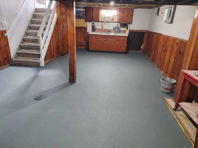 basement featuring wooden walls, stairway, wainscoting, electric panel, and a sink