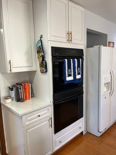 kitchen with white cabinets, black double oven, white refrigerator with ice dispenser, light stone countertops, and light wood-type flooring