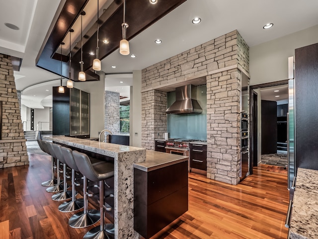 kitchen featuring light stone counters, wall chimney exhaust hood, decorative light fixtures, and a kitchen bar