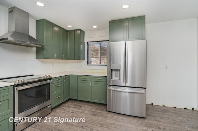 kitchen featuring stainless steel appliances, green cabinetry, wall chimney exhaust hood, and wood finished floors