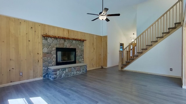 unfurnished living room with a stone fireplace, wooden walls, dark hardwood / wood-style flooring, ceiling fan, and a high ceiling