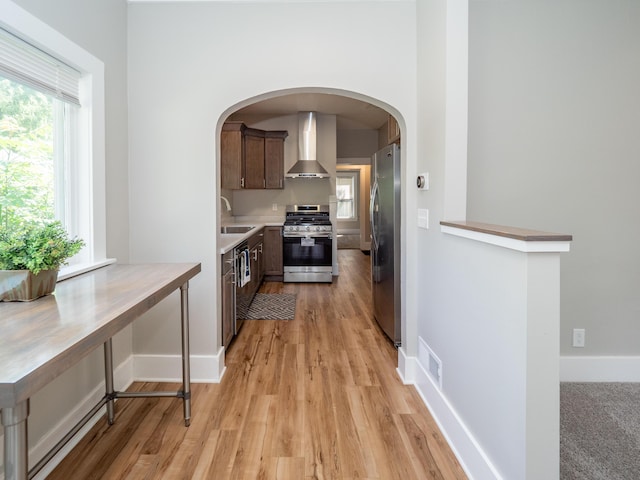 kitchen featuring wall chimney range hood, sink, appliances with stainless steel finishes, dark brown cabinets, and light hardwood / wood-style floors