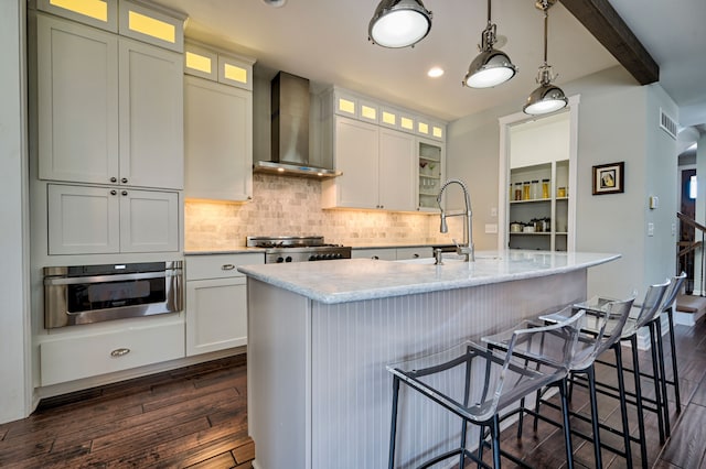 kitchen featuring wall chimney exhaust hood, decorative light fixtures, oven, and a kitchen island with sink