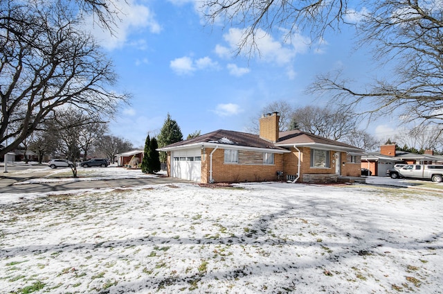 snow covered property featuring brick siding, an attached garage, a chimney, and driveway