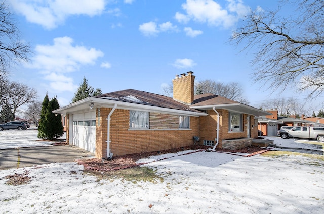 snow covered property with brick siding, driveway, a chimney, and a garage