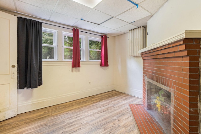 unfurnished living room featuring a drop ceiling, a brick fireplace, and light wood-type flooring